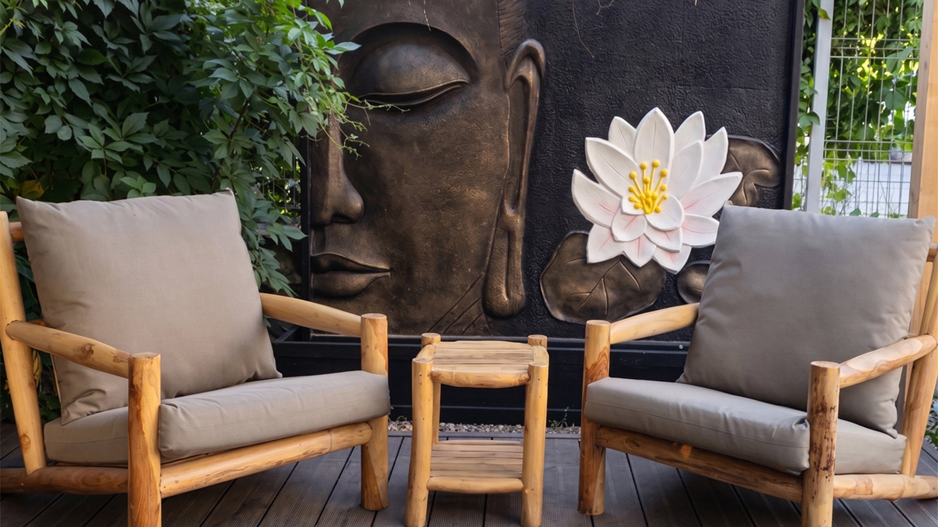 Mobilier de terasa din lemn de tec cu perne bej si masuta asortata, expus langa o fantana decorativa din fibrociment cu Buddha si floare de lotus, intr-un decor exterior cu vegetatie si fibre naturale.
